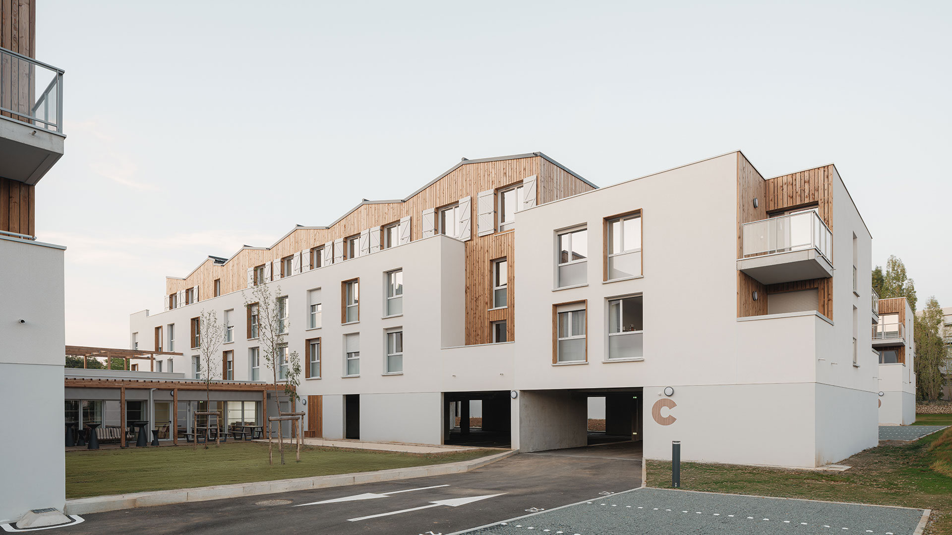 Vue extérieure d’une résidence contemporaine aux façades blanches et bardées de bois, avec espaces paysagers et terrasses aménagées.