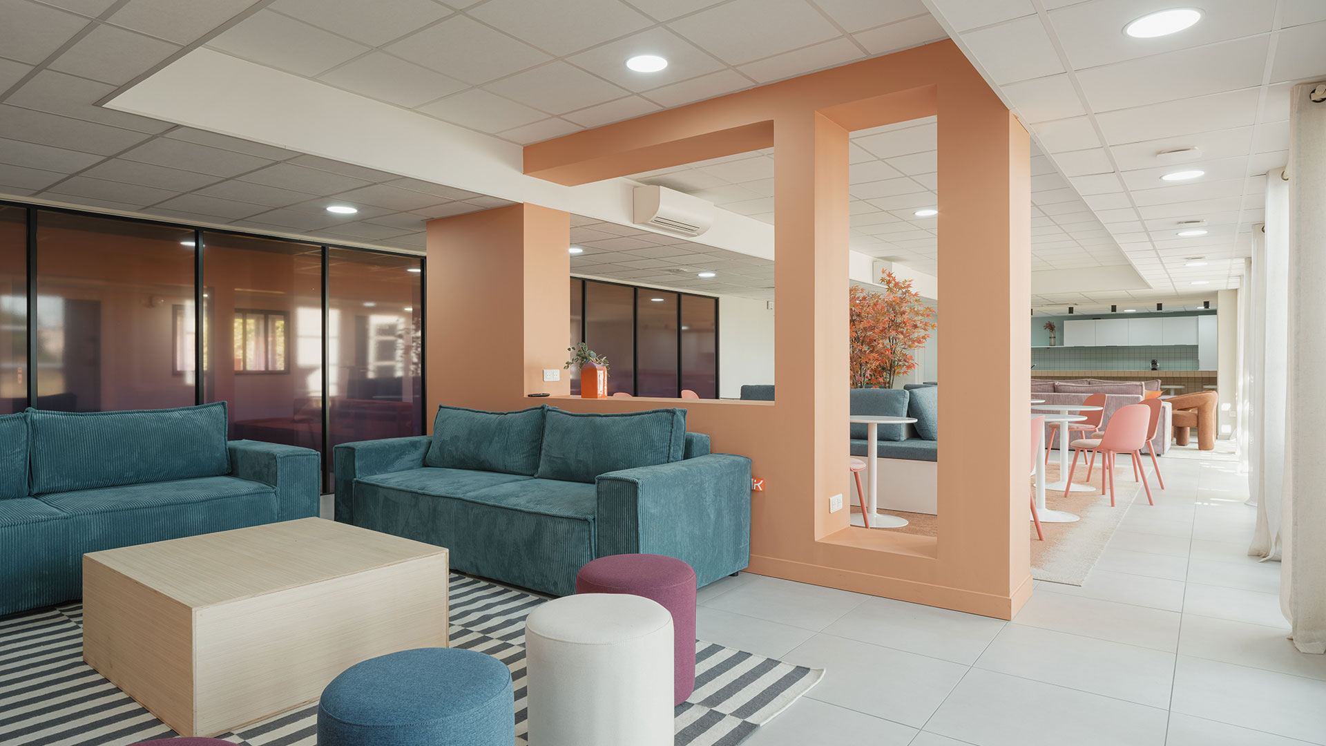Grande salle commune aménagée avec canapés, fauteuils et tables, ouverte sur un espace repas avec cuisine.