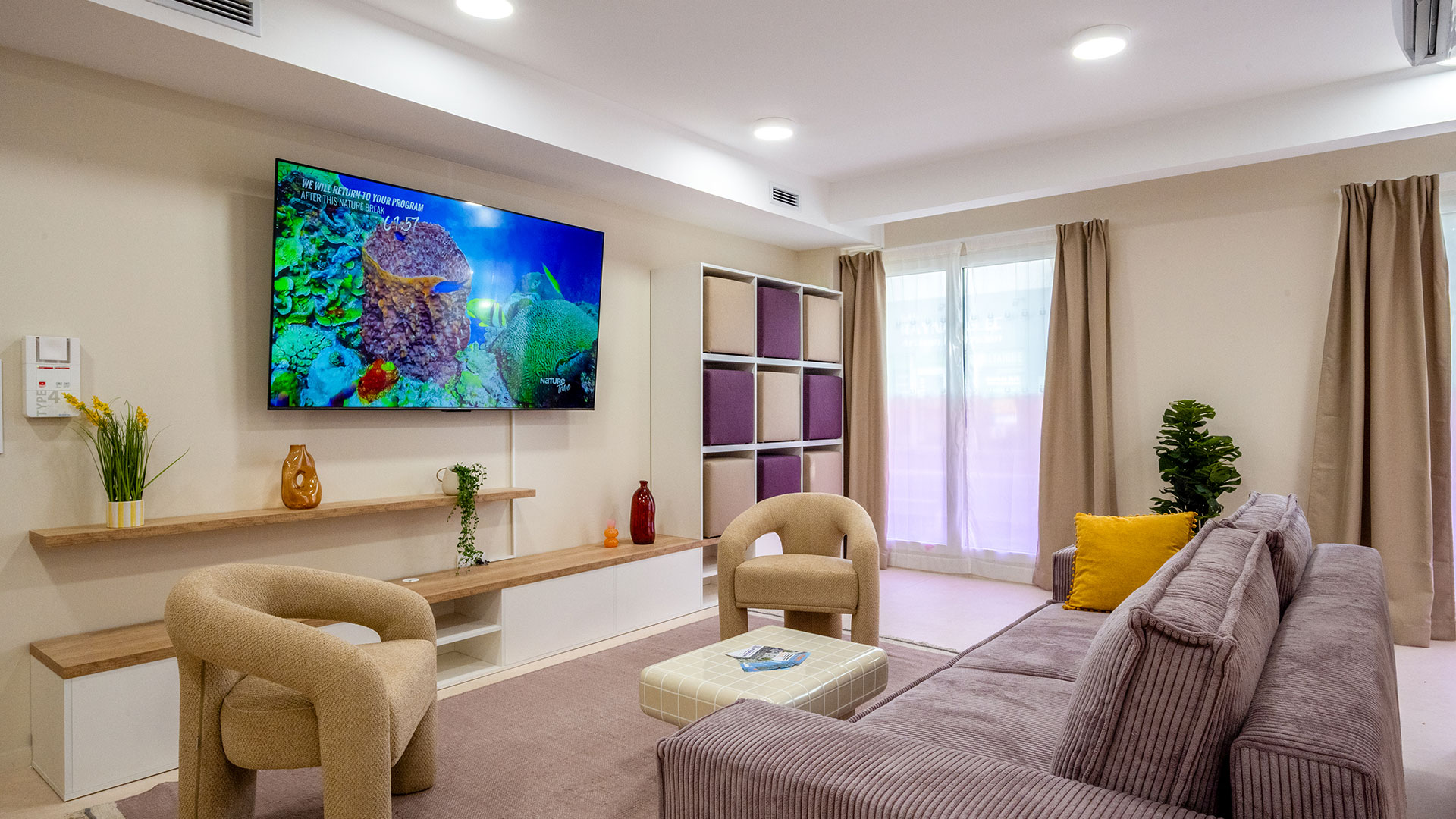 Salle de détente aménagée avec canapé, fauteuils, télévision et mobilier de rangement, baignée de lumière naturelle.